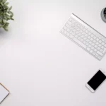 Mouse e teclado da Apple sobre uma mesa ao lado de vaso com plantas e celular