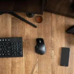 Teclado e mouse Dell ao lado de smartphone e controle de jogo em cima de uma mesa de madeira