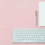 Teclado para escritório ao lado de mouse, caneta e caderno de notas sobre uma mesa com tampo rosa