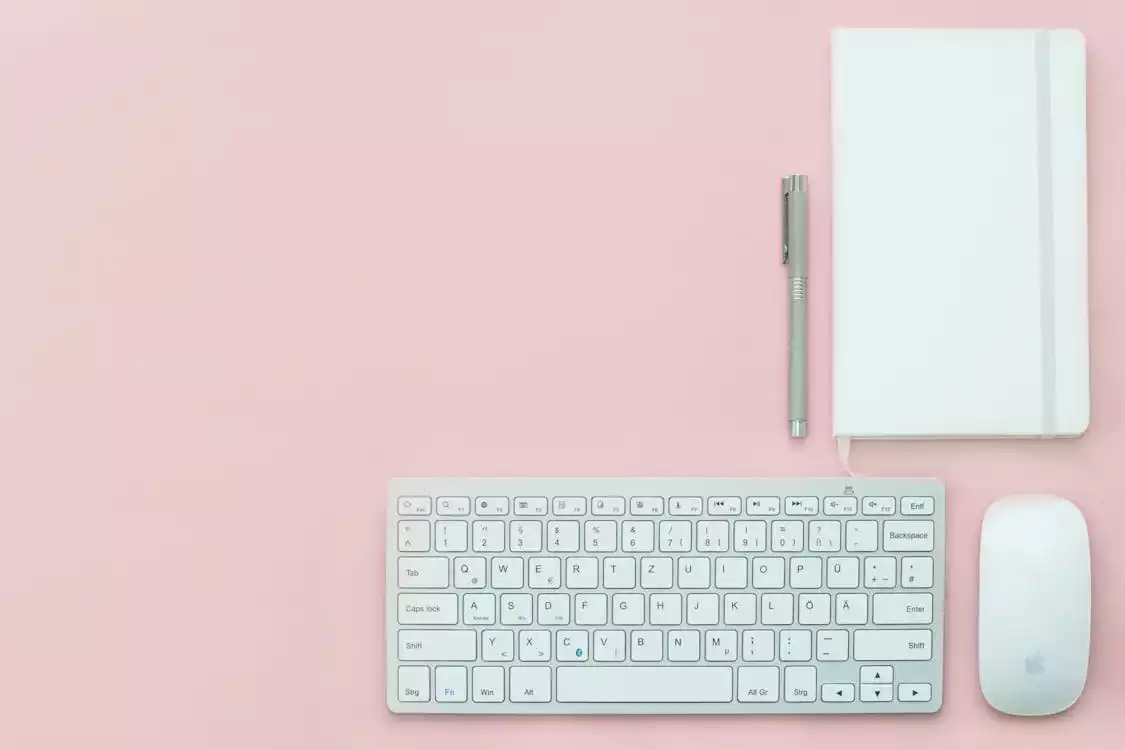 Teclado para escritório ao lado de mouse, caneta e caderno de notas sobre uma mesa com tampo rosa