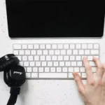 Pessoa usando teclado sem fio sobre uma mesa ao lado de um headfone e um tablet