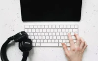 Pessoa usando teclado sem fio sobre uma mesa ao lado de um headfone e um tablet