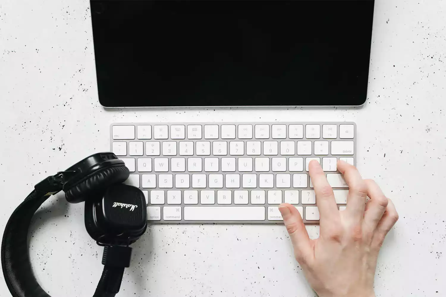 Pessoa usando teclado sem fio sobre uma mesa ao lado de um headfone e um tablet