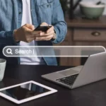 Moço buscando em site no celular serviço de dominio e hospedagem, atras de mesa com notebook e tablet e uma imagem de um buscador transparente