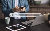 Moço buscando em site no celular serviço de dominio e hospedagem, atras de mesa com notebook e tablet e uma imagem de um buscador transparente