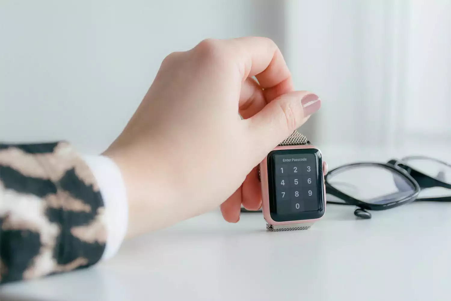 Moça segurando smartwatch em cima de uma mesa ao lado de um par de óculos