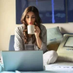 moça sentada na cama segurando uma xícara de café e usando um notebook barato