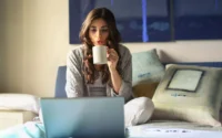 moça sentada na cama segurando uma xícara de café e usando um notebook barato
