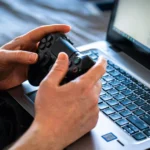 Mãos de jovem segurando controle joystick jogando em notebook gamer