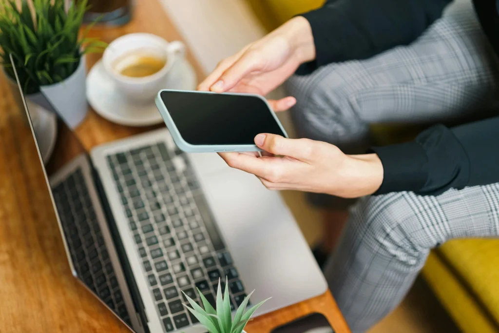 melhor-preço-de-celular-capa mulher segurando celular sentada em sofá em frente a notebook ilustrando como escolher o melhor preço de celular