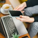 mulher segurando celular sentada em sofá em frente a notebook ilustrando como escolher o melhor preço de celular