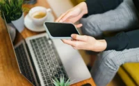 mulher segurando celular sentada em sofá em frente a notebook ilustrando como escolher o melhor preço de celular