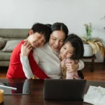 mãe abraçando dois filhos menino e menina sentada em frente a uma mesa com tablet, computador e desenhos em cima