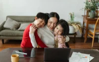 mãe abraçando dois filhos menino e menina sentada em frente a uma mesa com tablet, computador e desenhos em cima