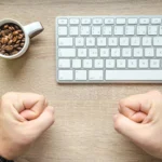 pessoa com as mãos fechadas como se estivesse torcendo sobre escrivaninha com teclado de computador ilustrando computador com bom custo-beneficio