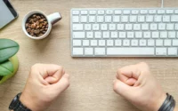 pessoa com as mãos fechadas como se estivesse torcendo sobre escrivaninha com teclado de computador ilustrando computador com bom custo-beneficio