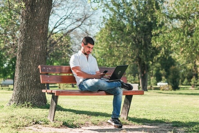 moço sentado em banco no parque segurando um notebook aberto sobre seu colo, ilustrando o assunto sobre notebook bom e barato